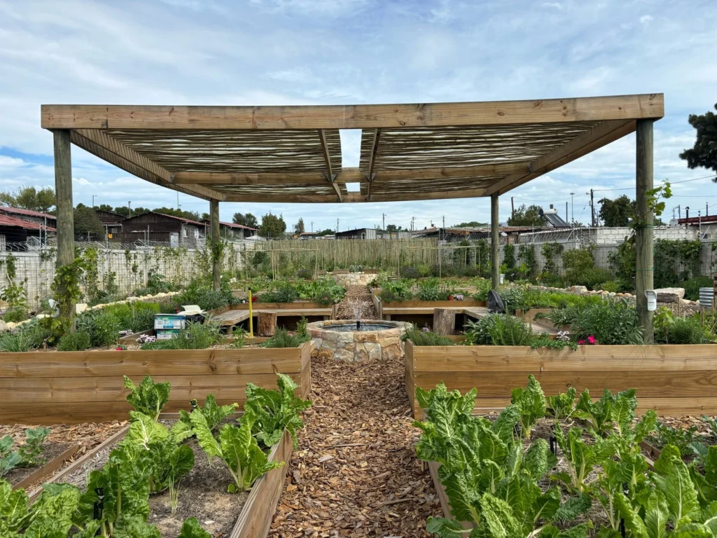 Holzpergola im Community Garden mit Hochbeeten, Sitzbänken und Brunnen als Treffpunkt im Freien.