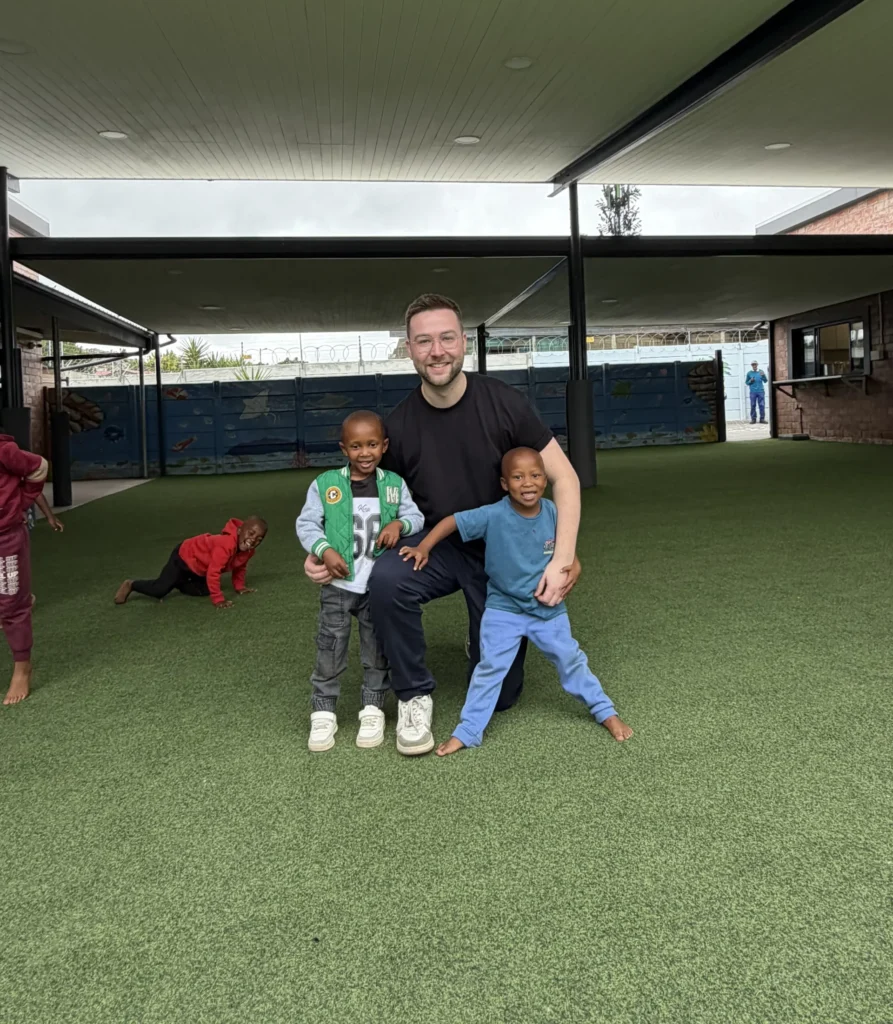 Betreuer kniet auf Kunstrasen und lächelt mit zwei Kindern in einem überdachten Innenhof.