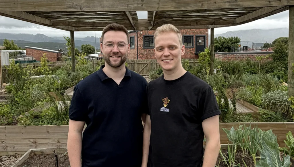 Zwei Teammitglieder im Gemeinschaftsgarten vor Hochbeeten unter einer Holzpergola.
